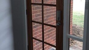Una puerta de madera con diseño de cuadrícula, que da a un espacio exterior con un muro de ladrillo y un césped.