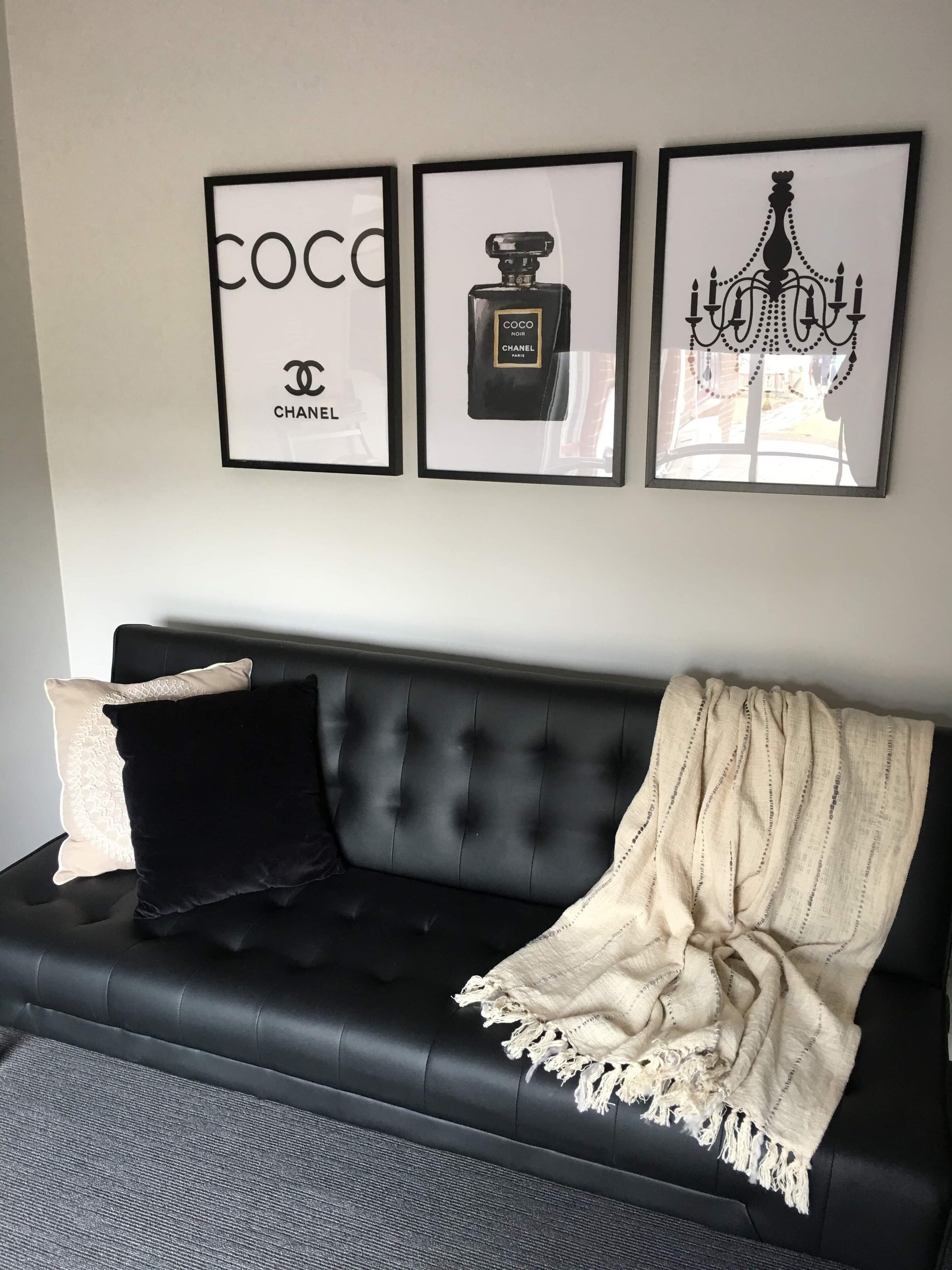 Un canapé noir agrémenté d’un plaid blanc et de deux coussins, un flacon de parfum Chanel encadré et un lustre encadré.