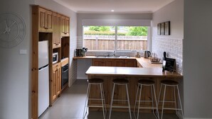 Una cocina moderna con armarios de madera, una isla central con taburetes, un ventanal con vista a un jardín trasero y un reloj de pared.