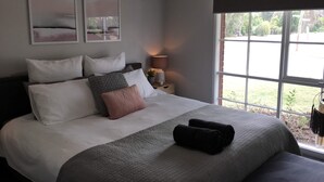Un dormitorio con una cama grande, varios almohadones, una lámpara de noche y un ventanal con vistas a la vegetación.