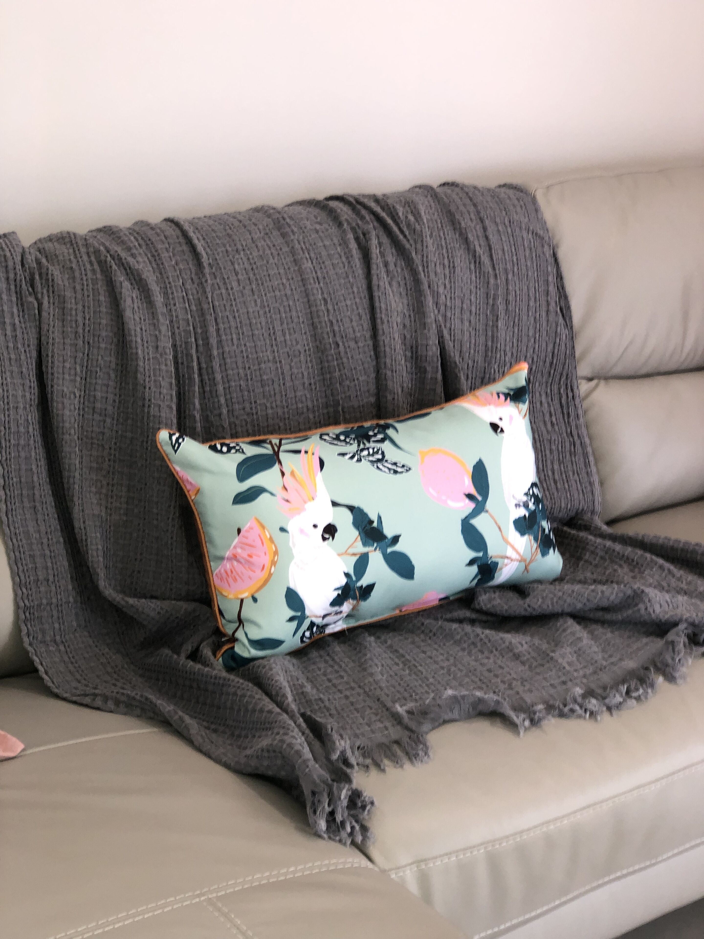 A sofa with a grey blanket and a cushion with a bird and floral pattern.