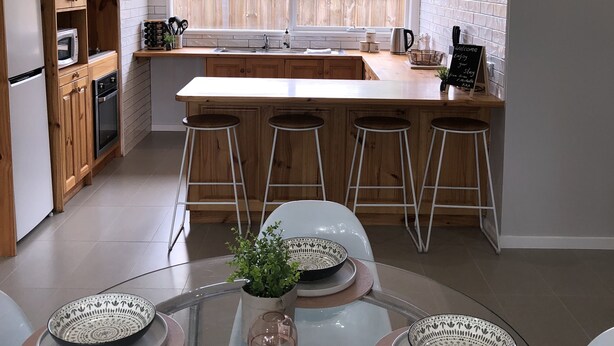 Una cocina moderna con armarios de madera, una isla grande con taburetes, una mesa de comedor de vidrio con cuencos estampados, y vistas a un patio trasero a través de amplios ventanales.