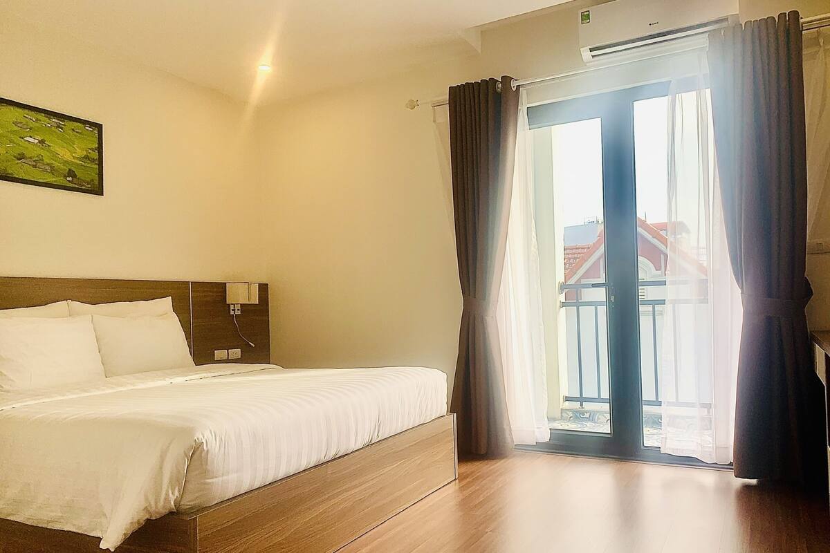 A hotel room with a bed, a nightstand, a wall-mounted lamp, and a view of the outside through a glass door.