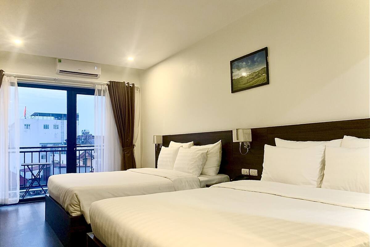 A hotel room with two beds, a large window with curtains, an air conditioning unit, and a painting on the wall.