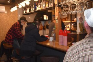 Zona de bar con taburetes, barra y estanterías con botellas y vasos.