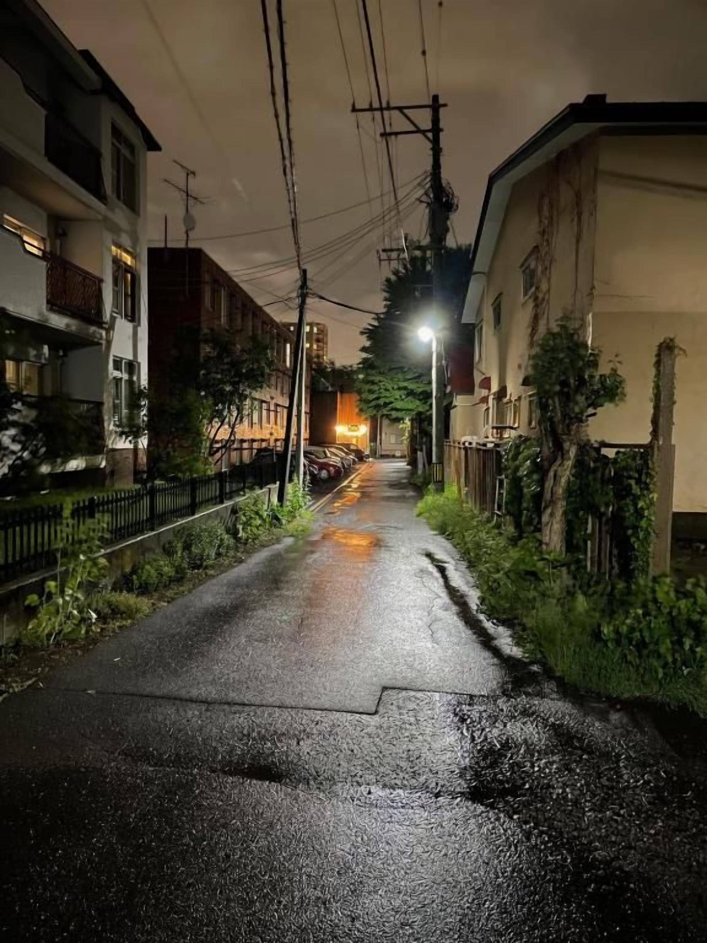 A narrow street with buildings on either side, a wet surface, and overhead power lines.
