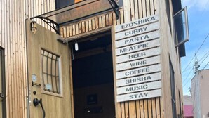 A wooden building with a sign listing amenities like curry, pasta, and coffee. A dog is lying on artificial grass in front. - Arura Sapporo - Hostel (Sapporo)
