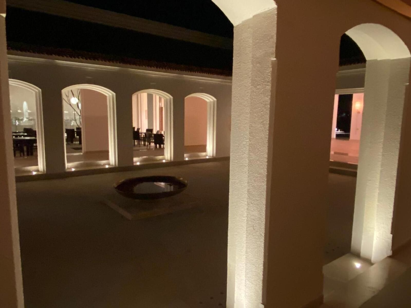 Un espacio al aire libre cubierto con una piscina, rodeado de pilares y arcos blancos, que conduce a un área interior con mesas y sillas.