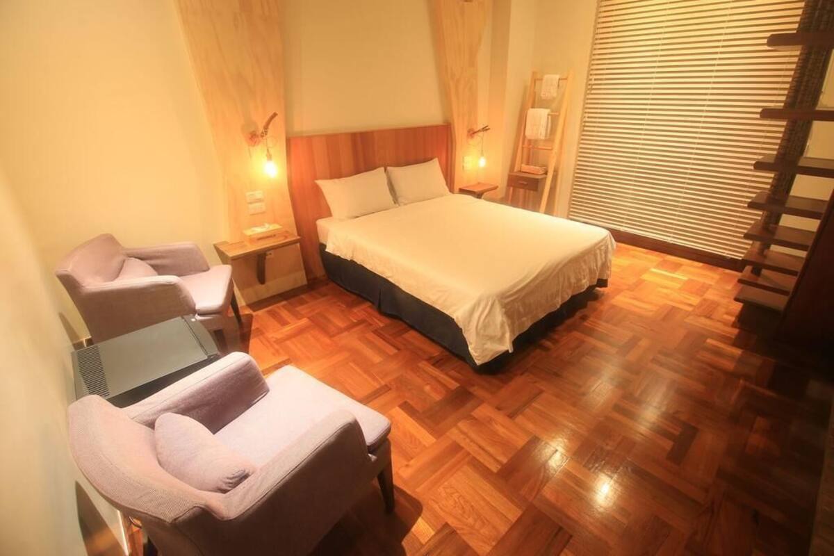 A hotel room with a bed, two armchairs, a small table, wooden flooring, and a window with blinds.
