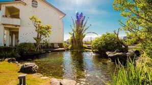 A serene garden with a pond, a white building with balconies, and lush greenery.