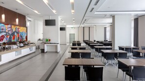 A modern dining area with long tables and black chairs, a buffet counter with various food items, and a colorful abstract painting on the wall. - Amaris Hotel Slipi (Jakarta)