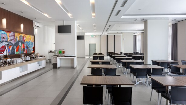 A modern dining area with long tables and black chairs, a buffet counter with various food items, and a colorful abstract painting on the wall.