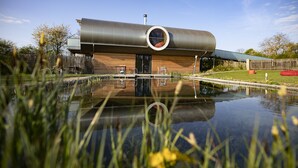 Un bâtiment moderne doté d’un toit cylindrique au design unique, d’un élément décoratif aquatique réfléchissant à l’avant et d’une terrasse en bois.