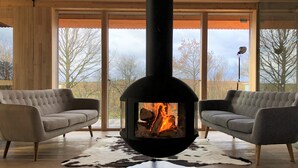 Un salon moderne doté d’un poêle à bois central, de deux canapés gris, d’un tapis en peau de vache et de grandes fenêtres offrant une vue sur les arbres et le ciel.