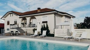 Une piscine avec des chaises longues et des parasols, un bâtiment blanc avec un toit en tuiles et un ciel dégagé.
