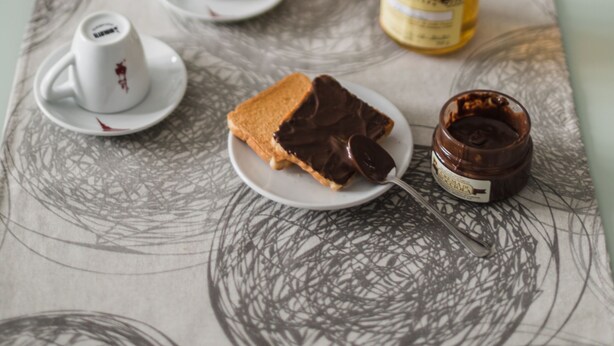 Une table recouverte d’une nappe à motifs, un panier de fruits, un pot de pâte à tartiner au chocolat, une cuillère et une tranche de pain garnie de chocolat.
