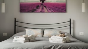 Un lit bien fait, avec des serviettes pliées, une tête de lit en métal et un tableau représentant un arbre dans un champ, accroché au mur.