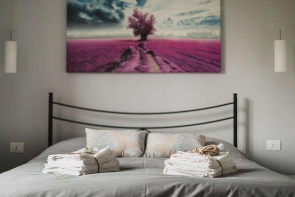 A neatly made bed with folded towels, a metal headboard, and a wall-mounted painting of a tree in a field.