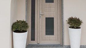 Une porte de couleur beige avec un panneau de verre, flanquée de deux jardinières blanches contenant des arbustes.