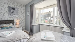 A bedroom with two beds, a nightstand with a lamp, a window with curtains, and a wall with a floral pattern and a painting. - Arkle House - a cosy home-from-home in Amble-by-the-Sea (Amble)