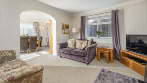 A living room with a sofa, a TV, and a window with curtains. - Arkle House - a cosy home-from-home in Amble-by-the-Sea (Amble)