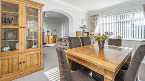 A dining area with a wooden table, cushioned chairs, a glass-fronted cabinet, and a window with curtains. - Arkle House - a cosy home-from-home in Amble-by-the-Sea (Amble)