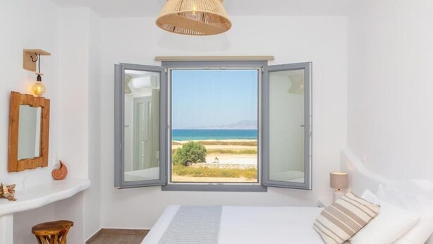 Un dormitorio con una cama grande, vista a la playa y una ventana con contraventanas abiertas.