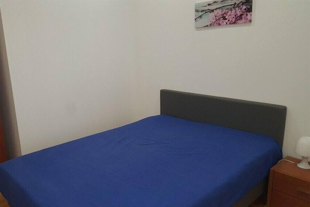 Una cama individual con una colcha azul, una mesita de noche de madera con una lámpara, y un armario de madera.
