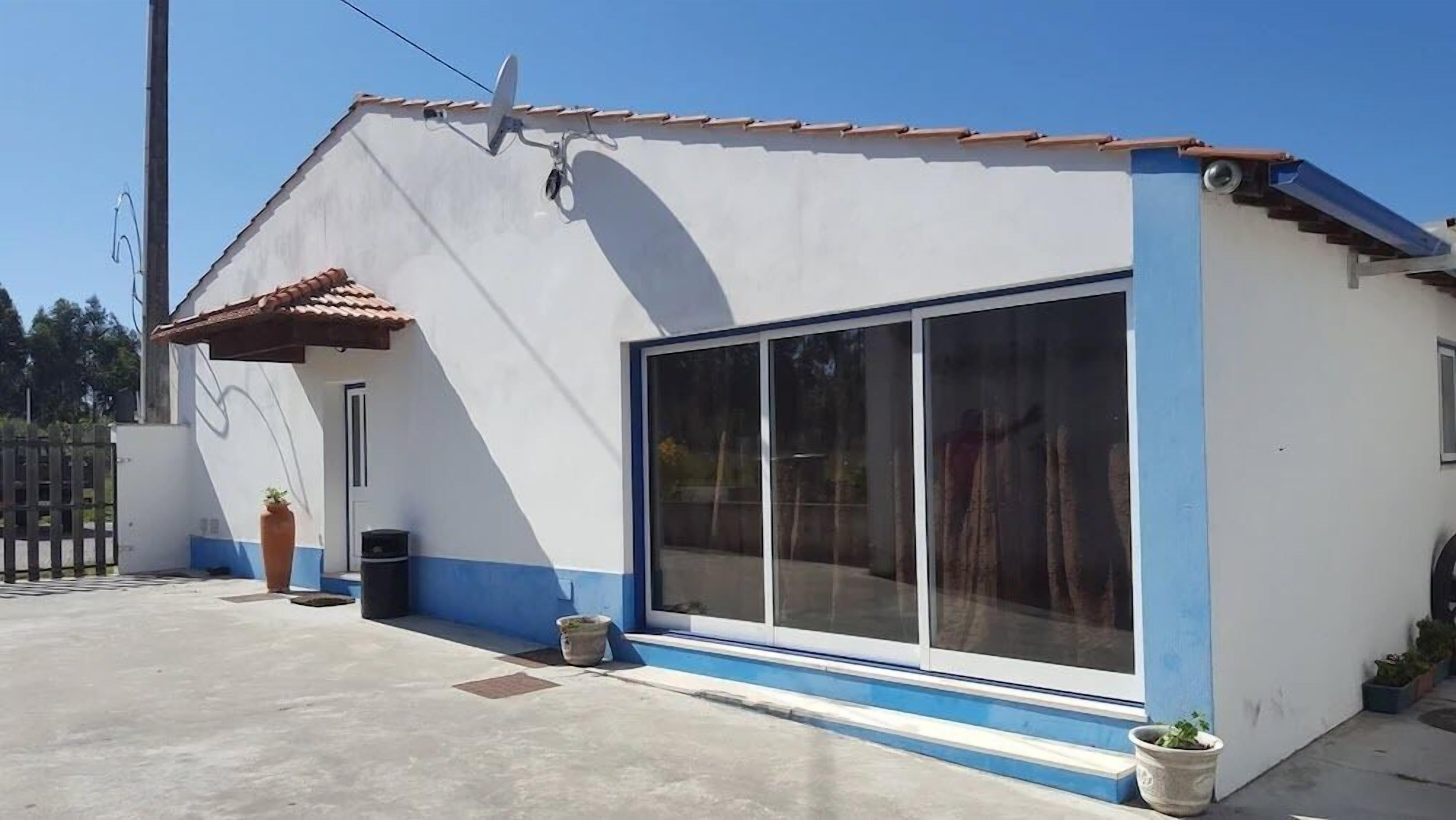 A small, single-story building with a white exterior and blue trim, featuring a large sliding glass door and a small porch area with potted plants.