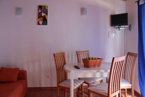 A room with a dining table, four wooden chairs, a red sofa, a television mounted on the wall, and a colorful abstract painting. - Quinta do Valão (Alcobaça)