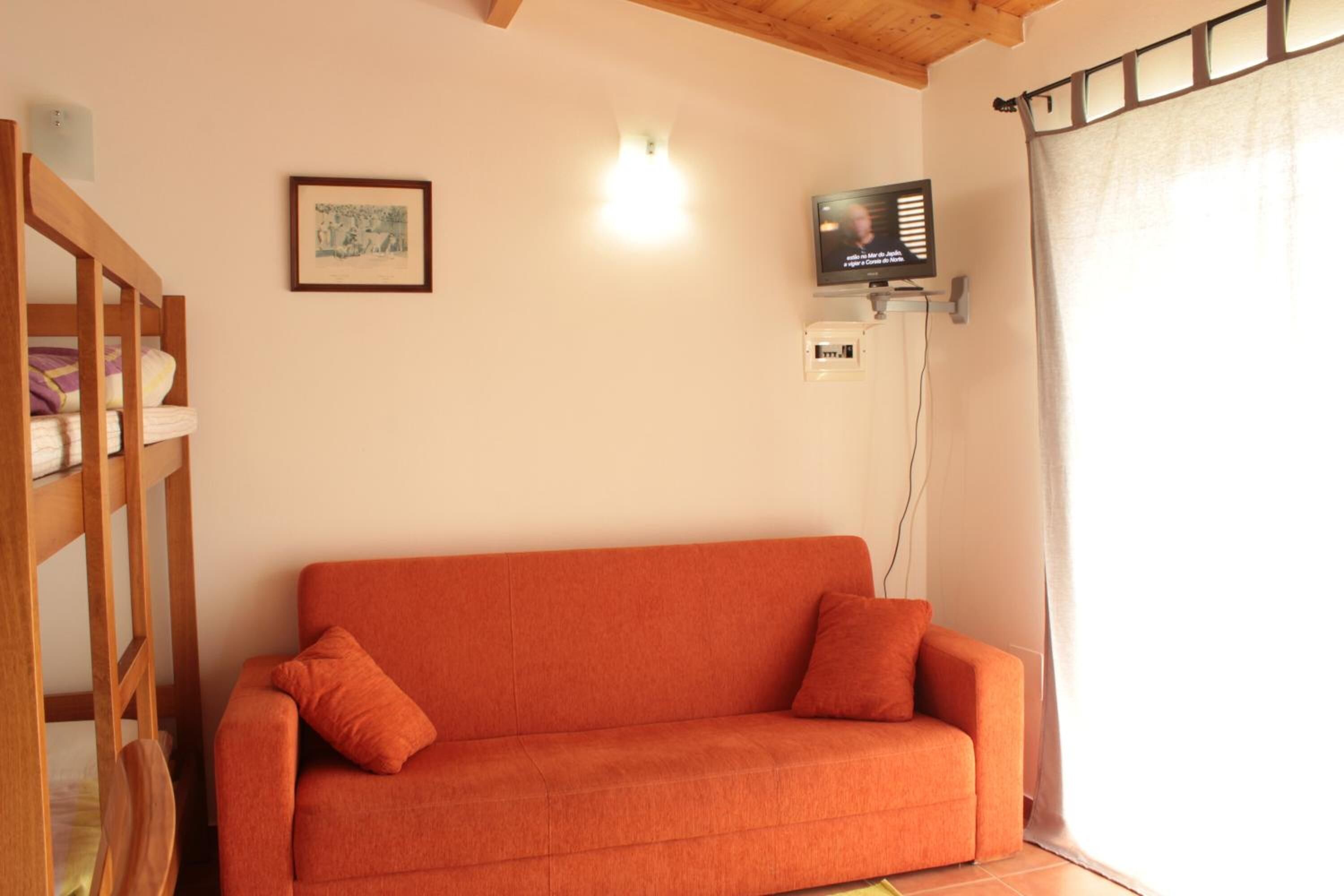 Ein Zimmer mit einem hölzernen Etagenbett, einem orangefarbenen Sofa, einem gerahmten Bild an der Wand, einem an der Wand befestigten Fernseher und einem Fenster mit Vorhängen.
