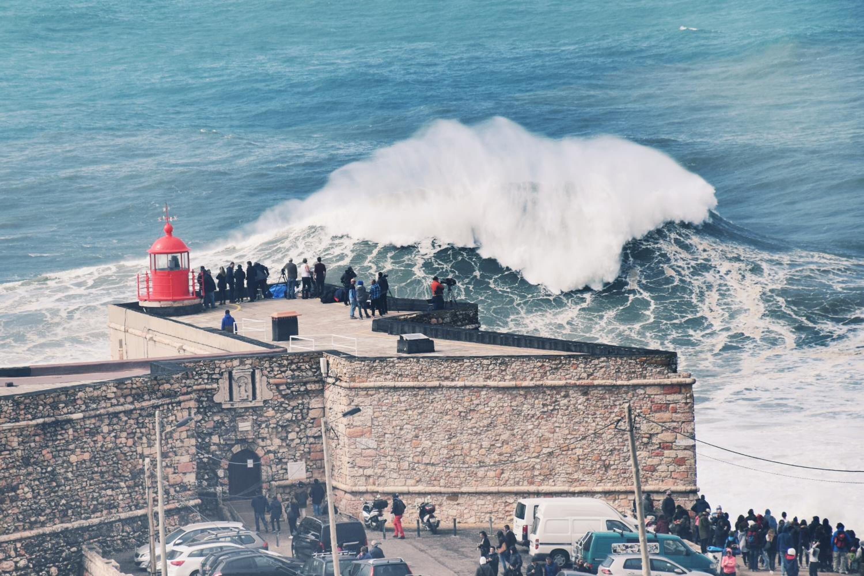A coastal scene with a stone building, a red lighthouse, and a large wave crashing onto the shore.