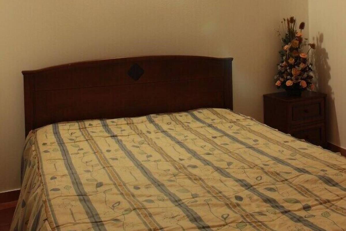 Un dormitorio con cabecera de madera, una cama individual con colcha estampada de flores, una mesita de noche con un florero y una lámpara de pared.