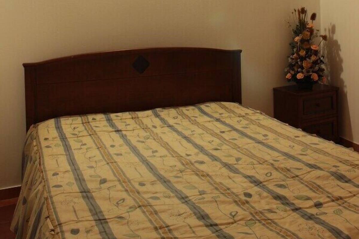 Un dormitorio con cabecera de madera, cama con colcha de estampado floral, mesita de noche con un jarrón de flores y lámpara de pared.