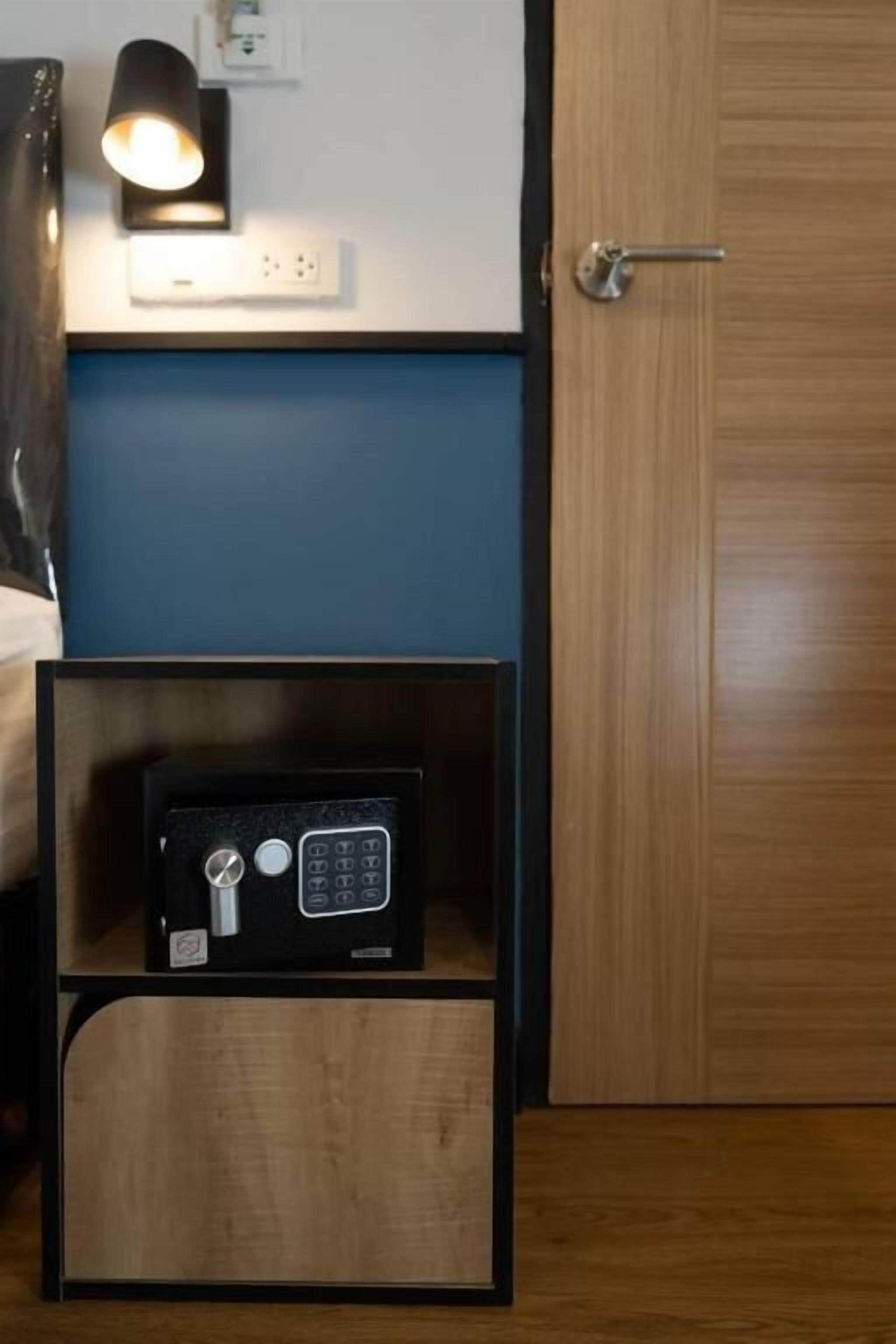 A room with a wooden door, a bedside table with a lamp, and a safe on the floor.