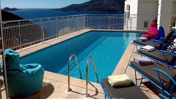 Ein Poolbereich mit Blick auf das Meer und die Berge, Liegestühle und ein weißes Gebäude.