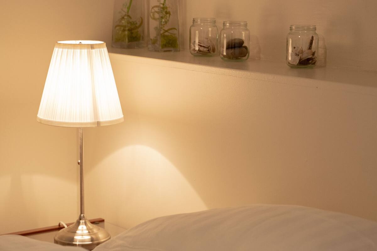 Ein Bett mit einem weißen Kopfkissen und einer Lampe auf einem Nachttisch. Darüber befindet sich eine Ablage mit drei Gläsern, gefüllt mit getrockneten Blumen und Blättern.