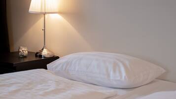 Een net opgemaakt bed met witte lakens en één wit kussen. Er staat een nachtkastje met een lamp en een glazen pot zichtbaar.