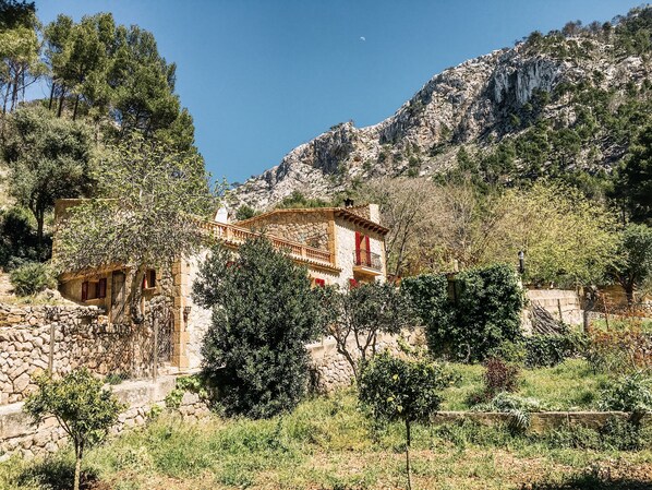 Edificio de piedra con balcón, rodeado de árboles y montañas al fondo.