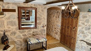 Una habitación rústica de paredes de piedra con una puerta de madera, un banco, un espejo y una araña.