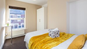 Een slaapkamer met een bed, een raam, een radiator, een witte kast en een kleintje tafeltje met een plant.
