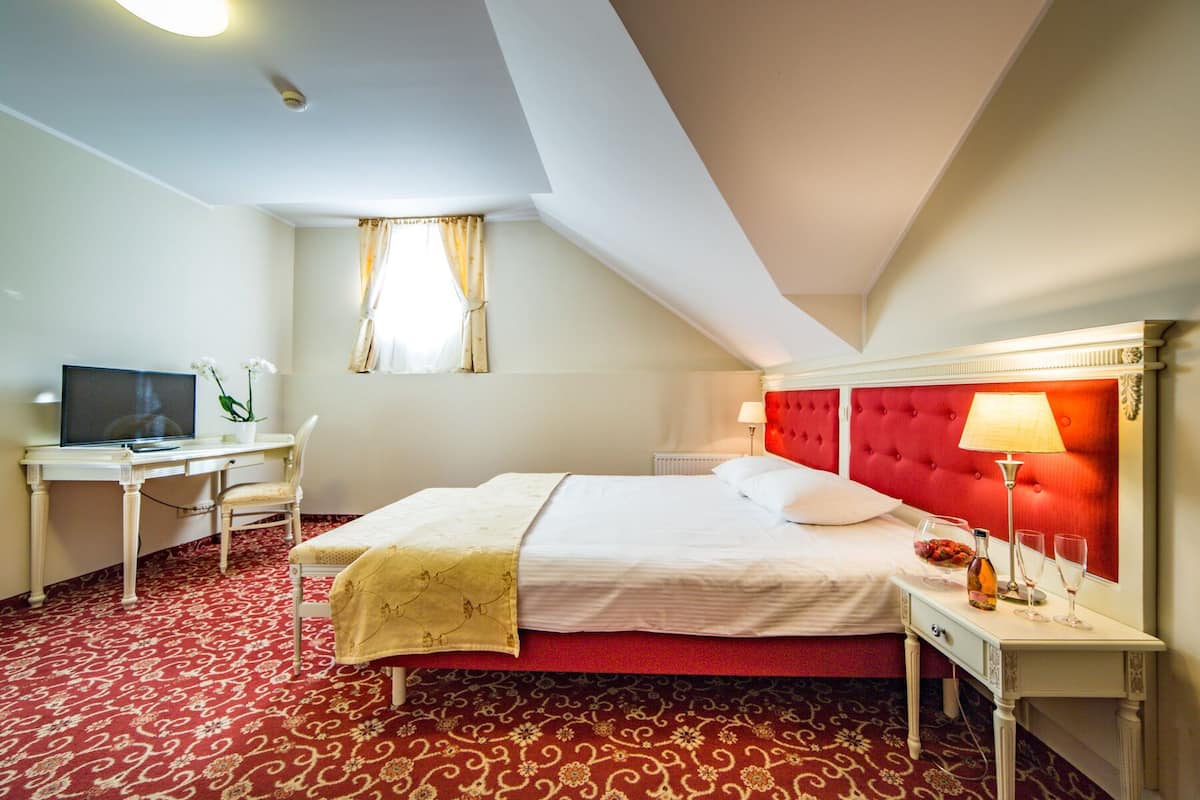 Quarto de hotel com uma cama, uma escrivaninha com computador, uma luminária e uma janela com cortinas.