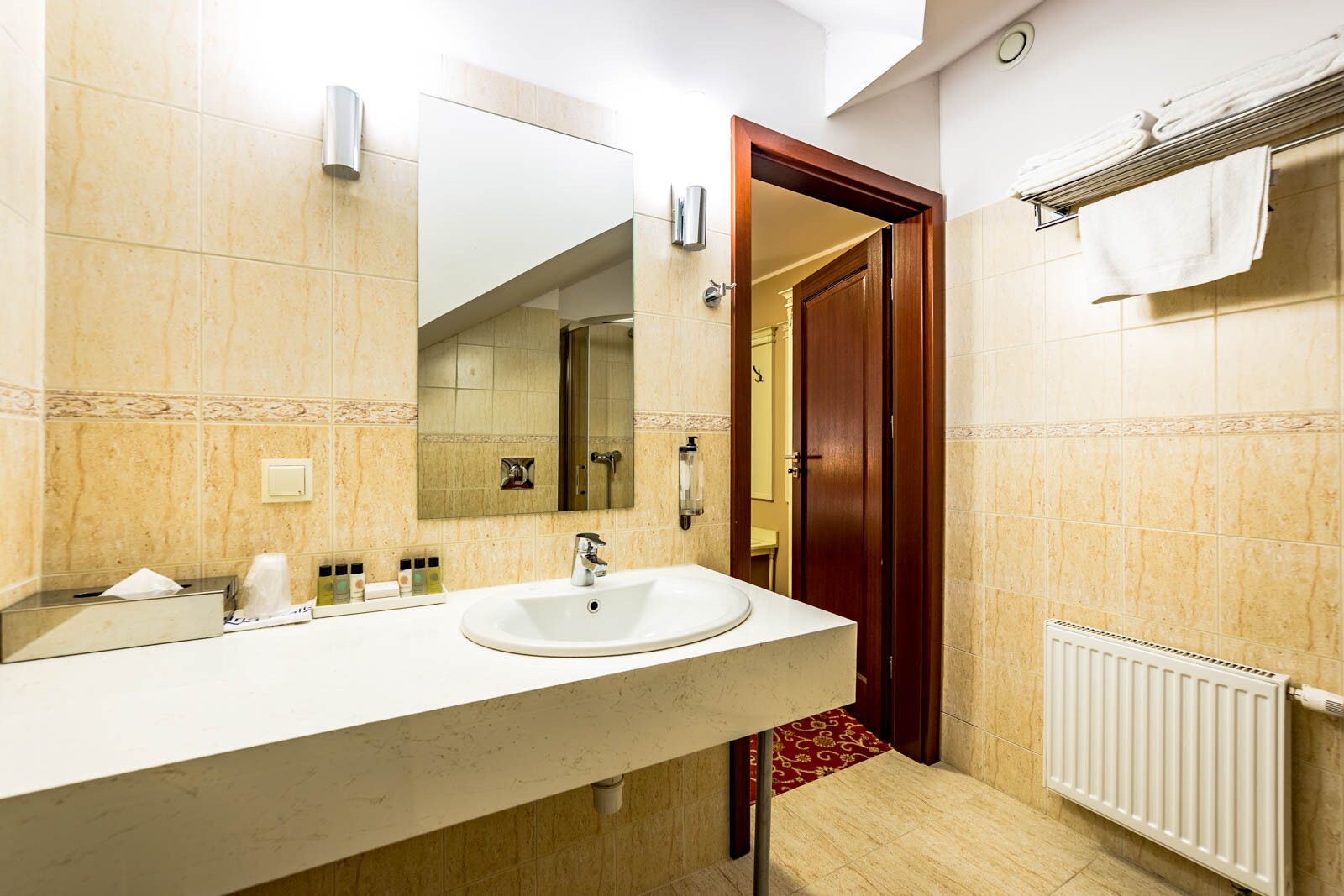 A hotel bathroom with a large mirror, a sink, a towel rack, and a radiator.