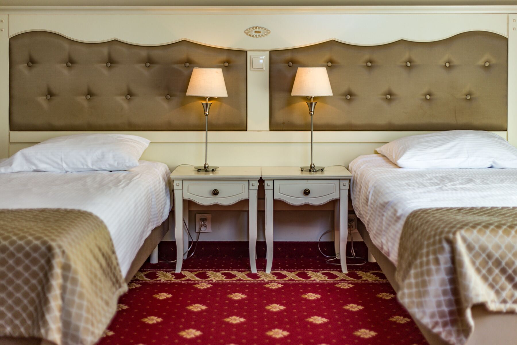 A hotel room with two beds, each with a white bedspread and matching pillows. There are two bedside tables with lamps, and a red patterned carpet.
