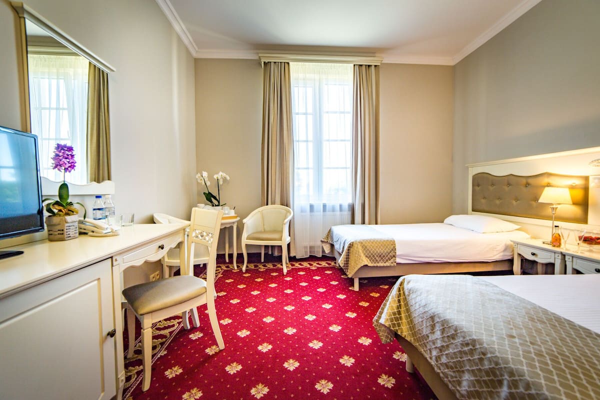 Quarto de hotel com escrivaninha, duas cadeiras, cama, televisão e janela com cortinas.
