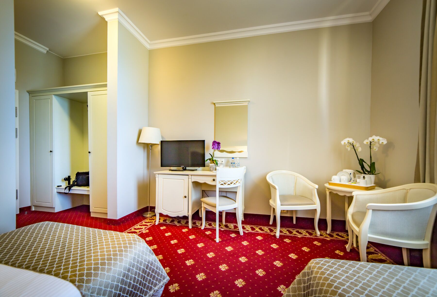 Una habitación de hotel con dos camas, un escritorio con computadora, una silla, una mesita con un florero y un espejo.
