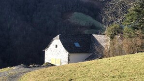 Un petit bâtiment en pierre, doté d’un toit en pente, d’une seule fenêtre et d’une porte, situé sur une colline herbeuse, avec une forêt en arrière-plan.