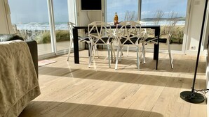 Ein Zimmer mit Holzboden, einem schwarzen Tisch mit weißen Stühlen, einem Fernseher an der Wand und Meerblick durch große Fenster.