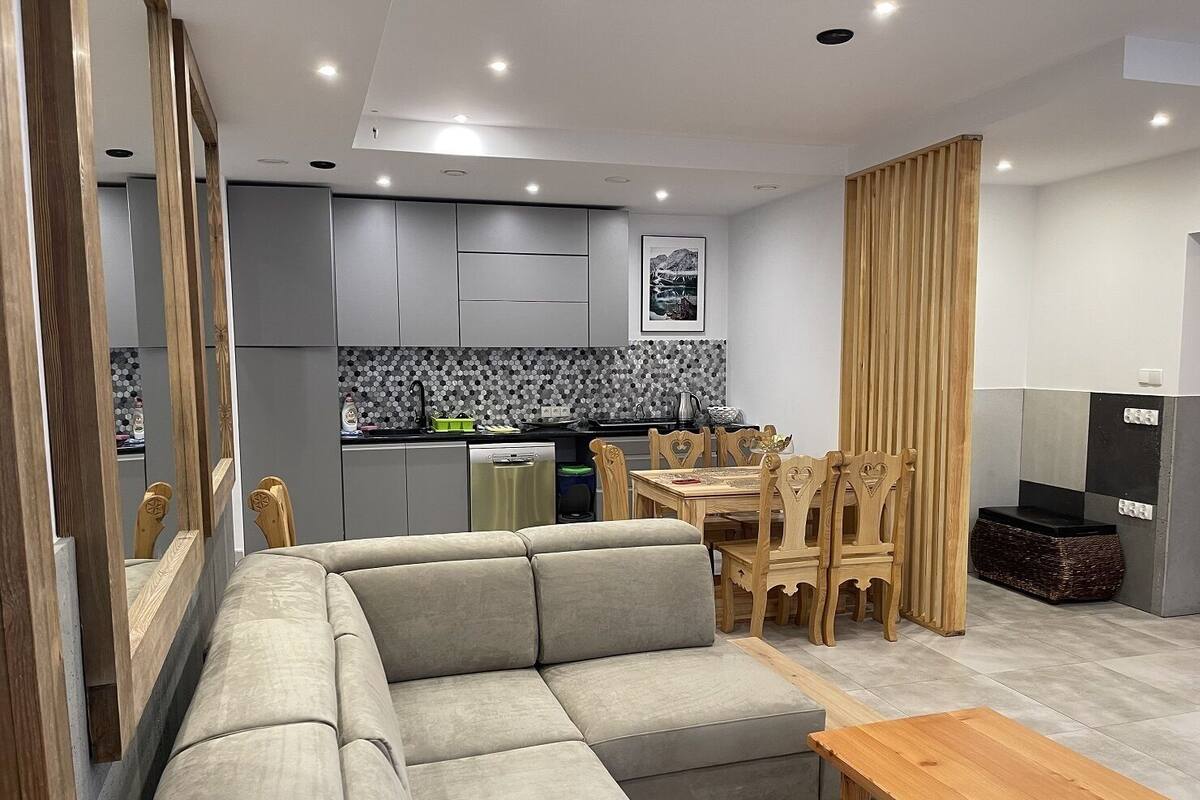 A modern living room with a grey sectional sofa, wooden dining table, and chairs. A kitchen with grey cabinets and a tiled backsplash is visible in the background.