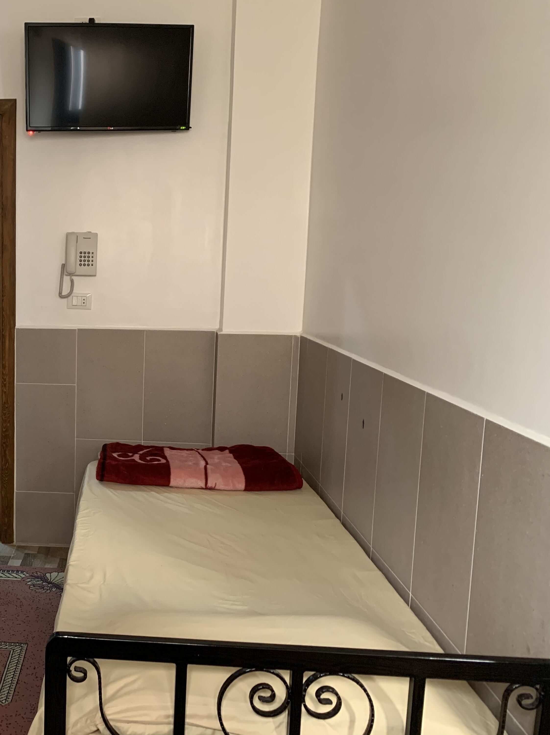 A single bed with a red and pink blanket, a mounted TV, a telephone, and a power outlet on the wall.